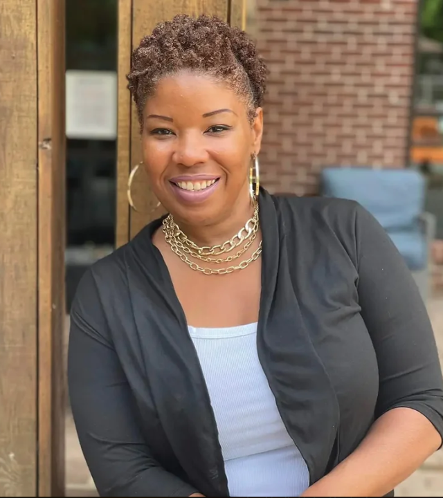 Smiling woman with braided hair wearing a layered necklace and black cardigan.